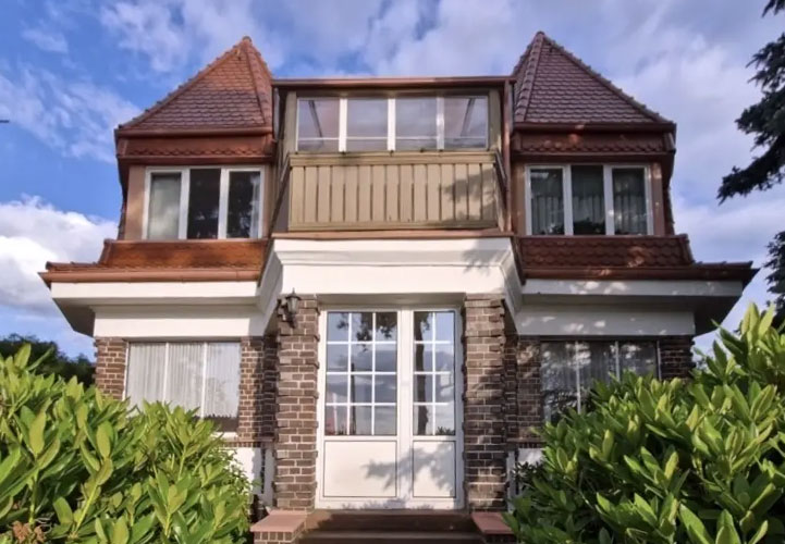 Eine elegante, moderne Villa aus hellem Backstein mit komplexer Dachlandschaft. Das zweistöckige Gebäude zeigt zeitgenössische Architektur mit verschiedenen Dacherker, einem markanten Mittelbalkon mit Glasgeländer und großen, weißen Sprossenfenstern. Die Fassade kombiniert hellen Klinker mit weißen architektonischen Akzenten. Eine breite Glastür mit weißen Rahmen bildet den Haupteingang. Das Haus ist von gepflegter Gartenlandschaft mit grünen Büschen und Pflanzen umgeben.
