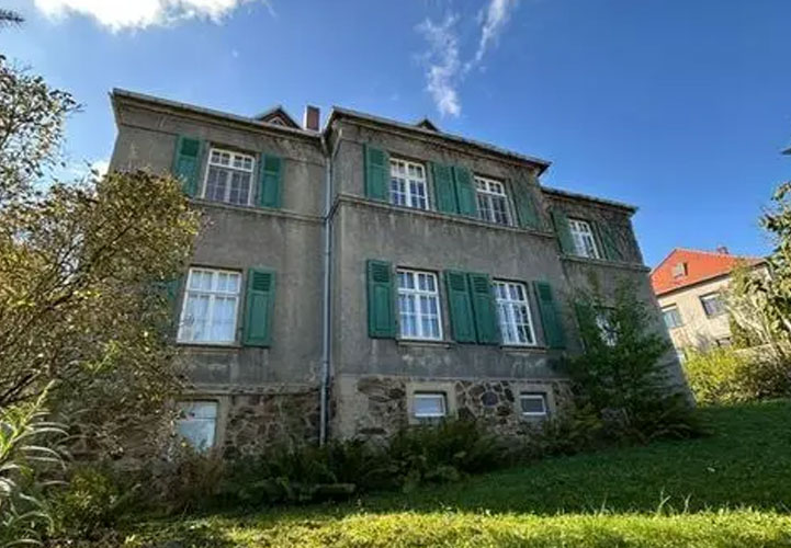 Eine zweistöckige Villa mit grauer Putzfassade und charakteristischen grünen Fensterläden an allen Fenstern. Das Gebäude hat ein rotes Ziegeldach und verschiedene architektonische Details. Im Erdgeschoss ist ein Natursteinsockel zu erkennen. Die Villa ist von einem verwilderten Garten umgeben mit hohem Gras und verschiedenen Pflanzen. Im Hintergrund sind weitere Gebäude mit roten Dächern zu sehen. Der blaue Himmel mit weißen Wolken und das natürliche Umfeld verleihen dem Anwesen einen rustikalen, ländlichen Charakter.