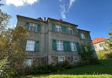 Eine zweistöckige Villa mit grauer Putzfassade und charakteristischen grünen Fensterläden an allen Fenstern. Das Gebäude hat ein rotes Ziegeldach und verschiedene architektonische Details. Im Erdgeschoss ist ein Natursteinsockel zu erkennen. Die Villa ist von einem verwilderten Garten umgeben mit hohem Gras und verschiedenen Pflanzen. Im Hintergrund sind weitere Gebäude mit roten Dächern zu sehen. Der blaue Himmel mit weißen Wolken und das natürliche Umfeld verleihen dem Anwesen einen rustikalen, ländlichen Charakter.