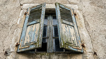 Doppelflügeliges Fenster mit stark verwitterten Holzläden an einer Steinmauer. Die Farbe blättert in Blau- und Weißtönen ab, die Fassade zeigt deutliche Risse. Algen- oder Moosbewuchs am unteren Rand der Läden.