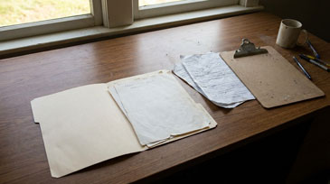 Holztisch vor Fenster mit einigen wenigen ausgebreiteten Papierdokumenten, Klemmbrett mit ein paar wenigen Notizen, und Kaffeetasse. Natürliches Tageslicht.