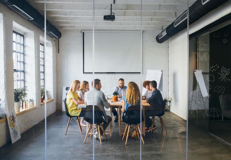 Welche Aufgaben erfüllt die Hausverwaltung? - wirtschaftliche, administrative Aufgaben Mehrere Personen sitzen an einem runden Tisch bei einem Meeting, man sieht sie durch eine Glasscheibe