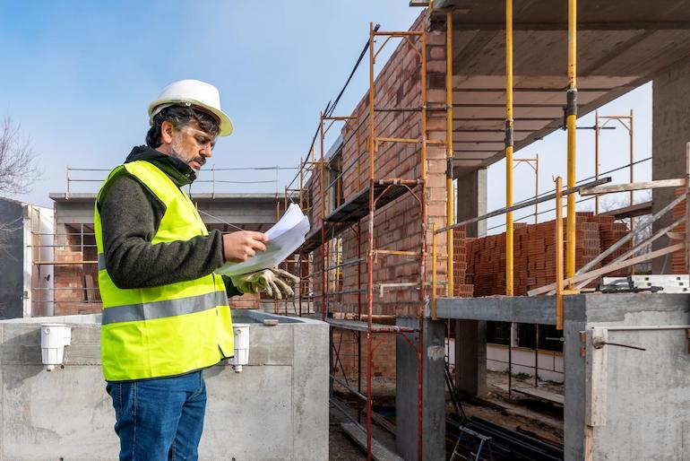 Welche Aufgaben erfüllt die Hausverwaltung? - Technische Aufgaben Mann mit Warnweste und Helm hat Klemmbrett in der Hand und steht vor einem Gebäude das saniert wird