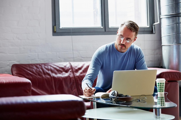 Mietvertrag kündigen wegen Zahlungsverzug Vermieter mit Lesebrille sitzt auf dem Sofa am Laptop, er notiert in einen Block Zahlen.